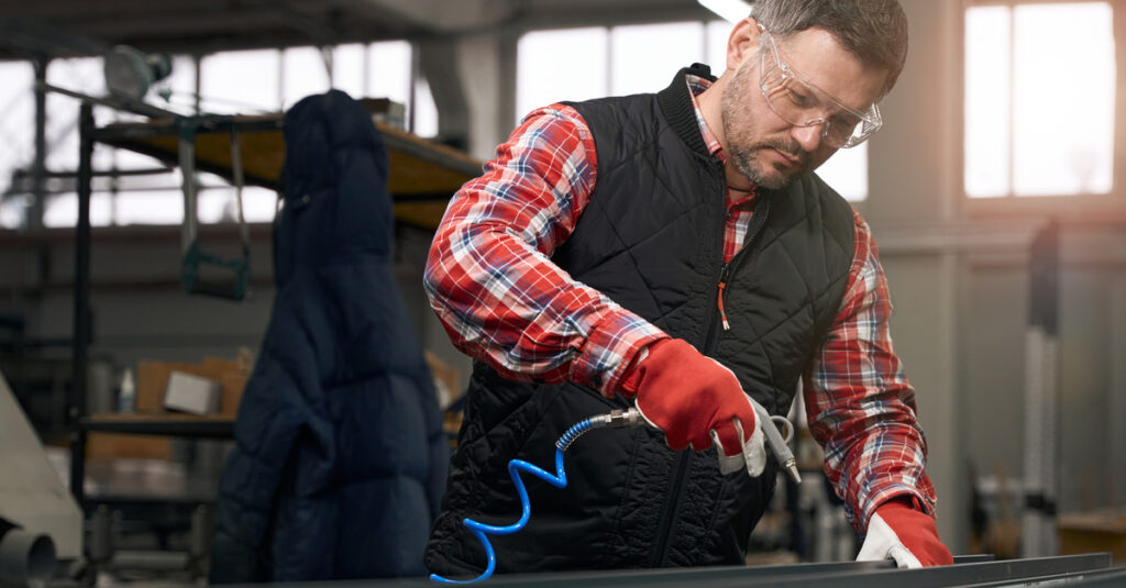 A man wearing a flannel shirt and vest with eye protection using a small air gun attached to a compressor.