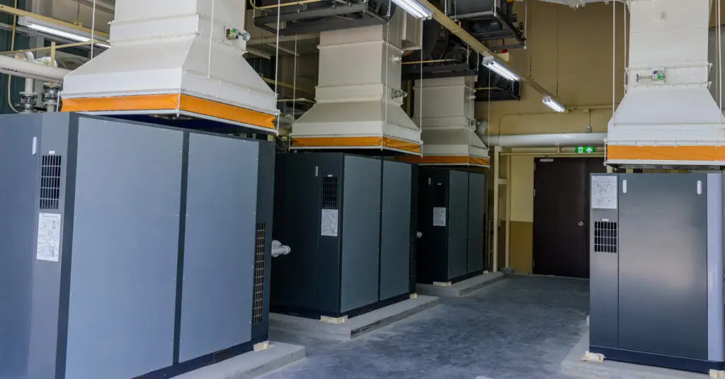 A group of industrial air compressors sitting on slabs over a cement floor attached to HVAC ducts in a factory.