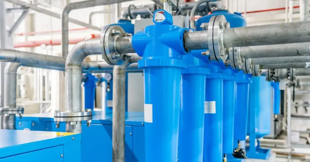 A blue air compression system in a warehouse attached to various metal pipes with a pressure gauge on the top.