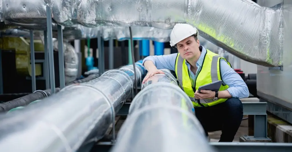 Un hombre con un chaleco amarillo de alta visibilidad y un casco blanco inspecciona unos tubos plateados de HVAC. - US Air Compressor A man wearing a yellow high-visibility vest and a white hard hat inspects some silver HVAC tubing.
