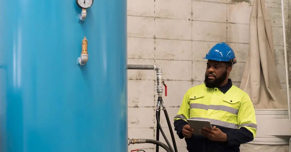 A bearded man in a high-visibility jacket and blue hard hat holds a clipboard and looks at a blue air compressor.