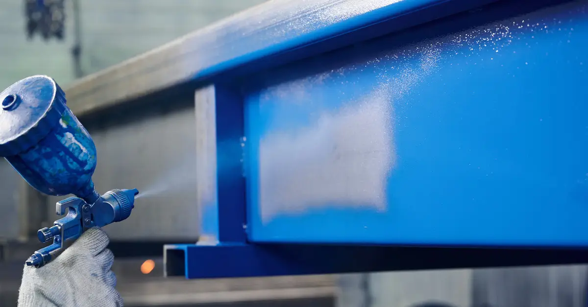 A hand wearing a white glove uses an air brush to spray paint a large, metal item blue.