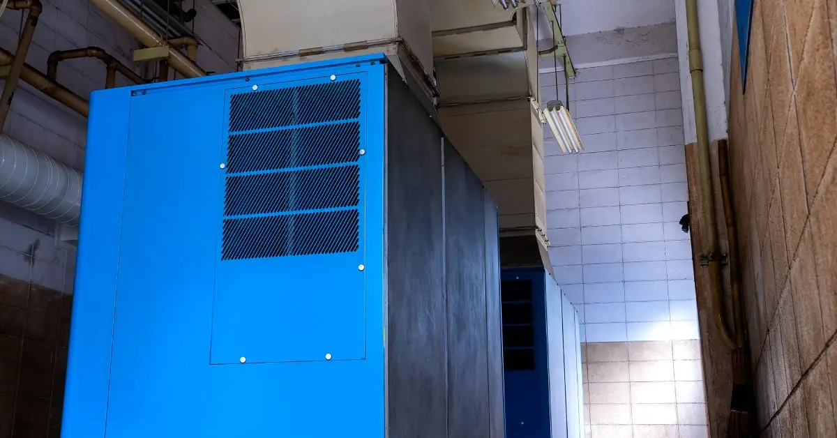A large, blue industrial air compressor unit set up in a factory with tile walls and low lighting.