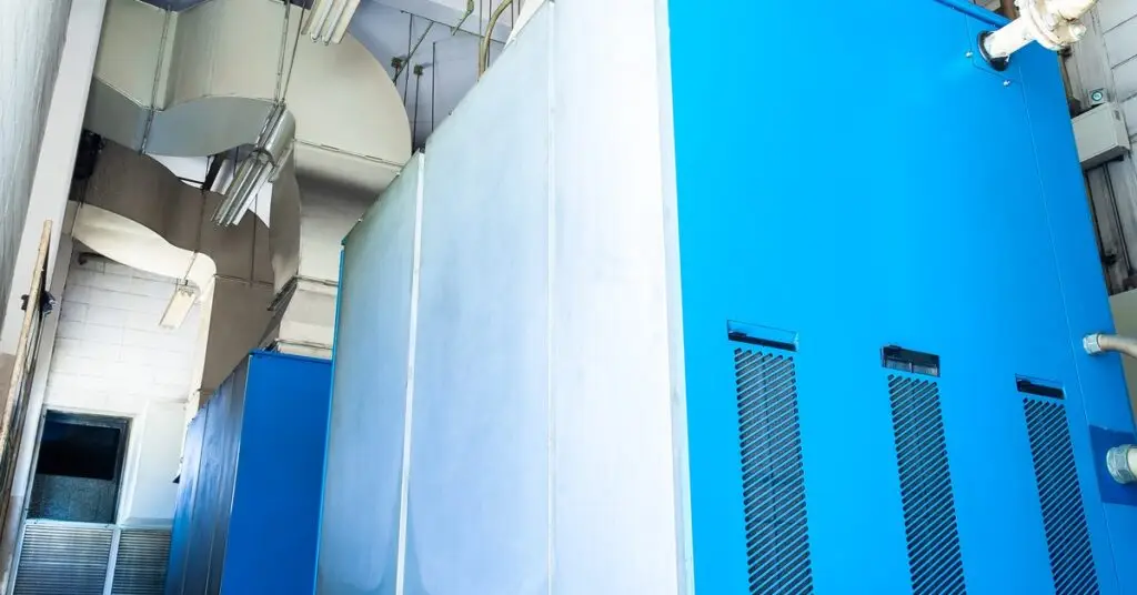 A large, light blue industrial air compressor in a dimly lit factory with white walls and HVAC vents. - US Air Compressor A large, light blue industrial air compressor in a dimly lit factory with white walls and HVAC vents.