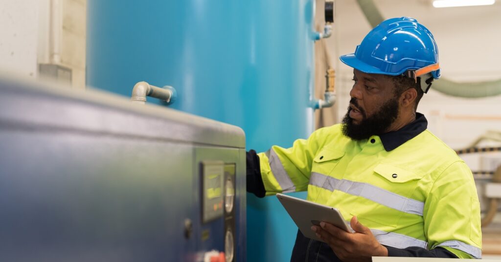 A bearded man in a high-visibility vest and hard hat inspecting an air compressor system's air tank.
