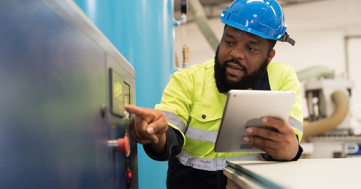 A bearded man in a high-visibility vest and hard hat pointing at a screen on an air compressor system.
