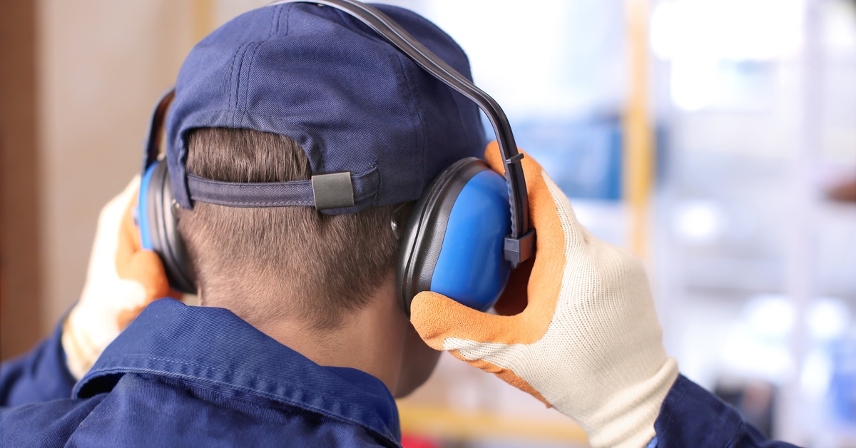 A person in a blue collared jumpsuit and a baseball hat wearing gloves puts on over-the-ear hearing protection.