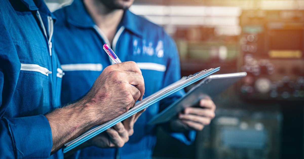 Two people wearing blue jumpsuits with white accents on them write on clipboards in an industrial setting.