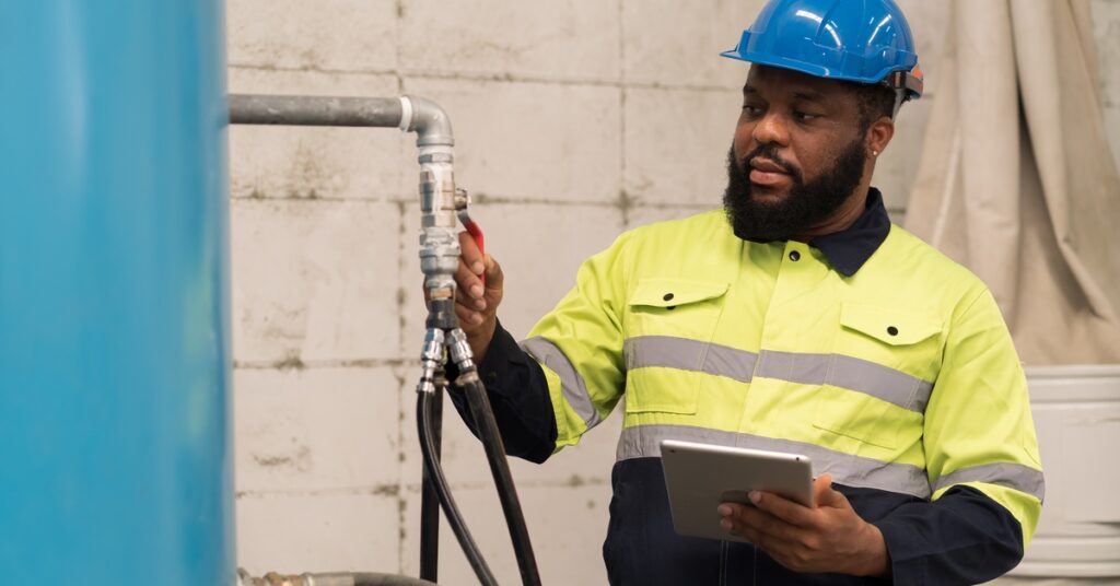 A man in a yellow and black high-visibility jacket and blue hard hat checks the pipes and tubing of an air compressor.