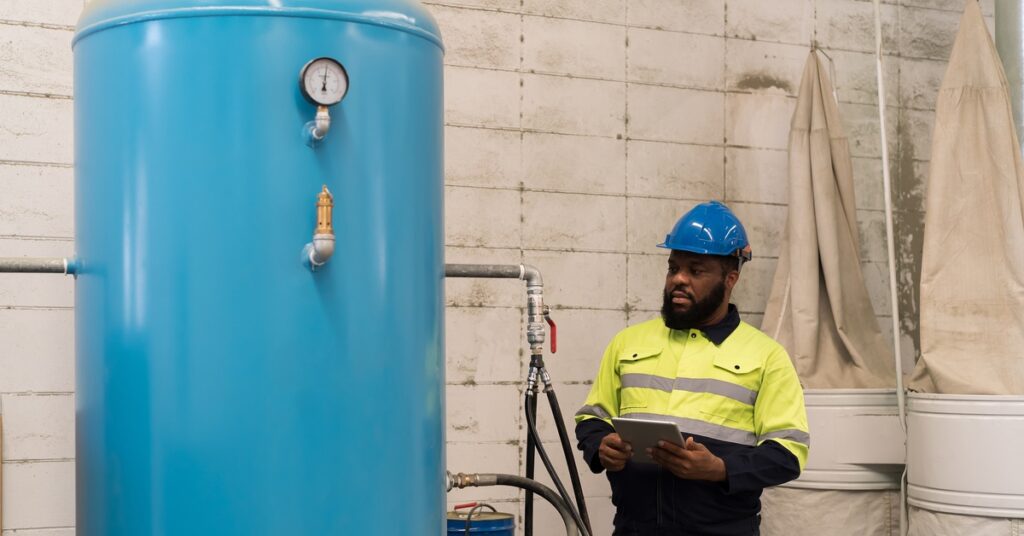 Un hombre con una chaqueta de alta visibilidad amarilla y negra sostiene un iPad y mira un compresor de aire azul claro. - US Air Compressor A man in a yellow and black high-visibility jacket holds an ipad and looks at a light blue air compressor machine.