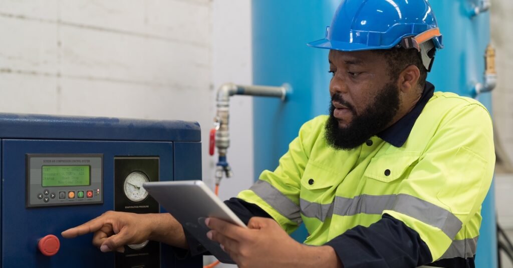 A man in a yellow and black jacket and blue hard hat points at an air compressor system while holding an ipad.