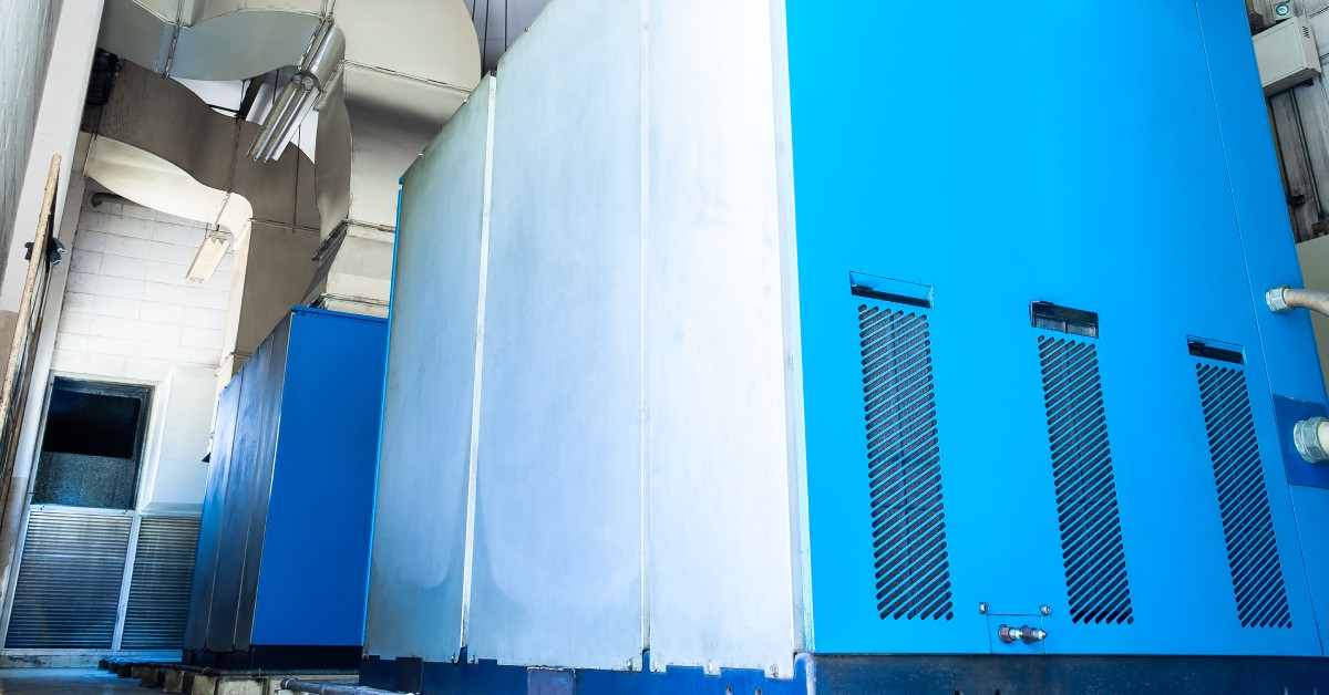 Two large blue and white air compressors with silver HVAC vents leading out of them in a low-lit facility.