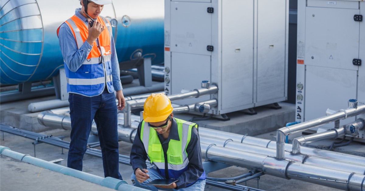 Two people wearing high-visibility vests, hard hats, and goggles performing HVAC maintenance on a roof.