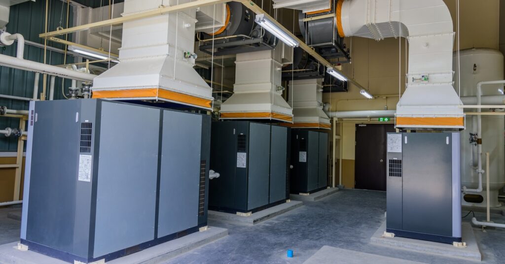 Four gray air compressor systems with large white and orange HVAC vents extending from them in a room. - US Air Compressor Four gray air compressor systems with large white and orange HVAC vents extending from them in a room.