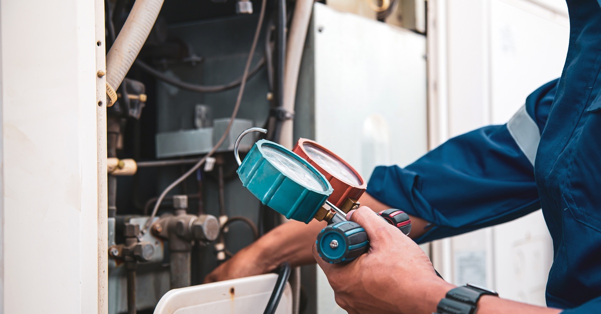 An HVAC technician in a blue jumpsuit holding a blue and red air gauge performing maintenance on an air compressor system.