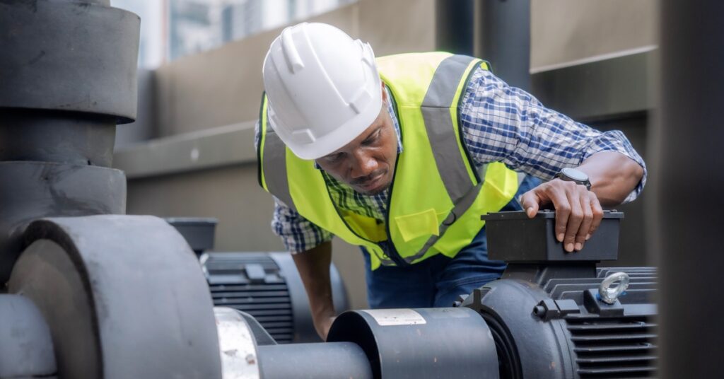 A man in a yellow high-visibility vest and a white hard hat inspects an air compressor unit on a roof.